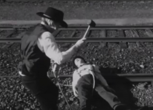 A screenshot from the film, showing a man tied to railroad tracks, and a villain threatening him with a hammer.